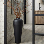 Tall black dimpled vase styled with twisting bare branches on a wood-look floor beside a pale brick wall and large black-framed mirror.
