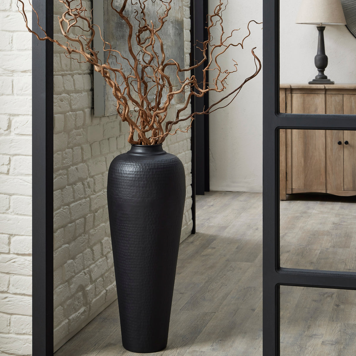 Tall black dimpled vase styled with twisting bare branches on a wood-look floor beside a pale brick wall and large black-framed mirror.