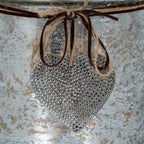 Close-up of the silver-toned heart pendant with shallow dimples, suspended beneath loops of twine and dark cord.