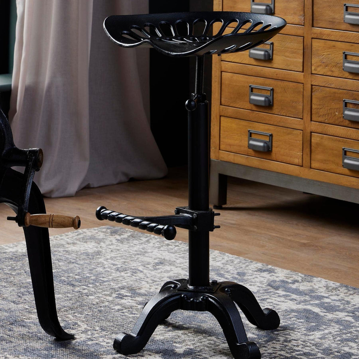 Dished Seat Stool standing on a patterned rug beside wooden drawers, with the central column and twisted footrest in view.
