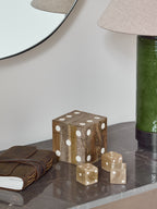 Closed dice box and three wooden dice on a dark veined tabletop beside a leather-bound book and green lamp.