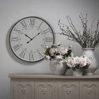 Doublehour Dial Wall Clock on a grey wall above a carved sideboard, styled beside pale vases holding flowers and tall branches.