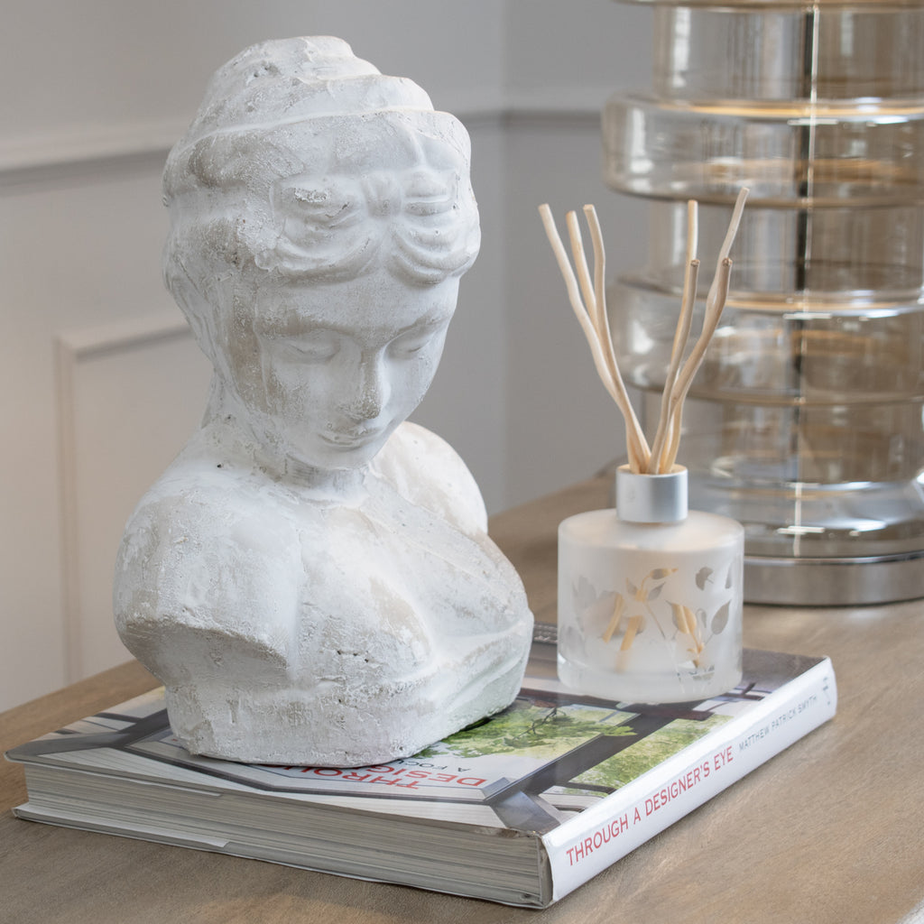 Pale female bust displayed on stacked books on a wooden console beside a reed diffuser and a glass lamp.