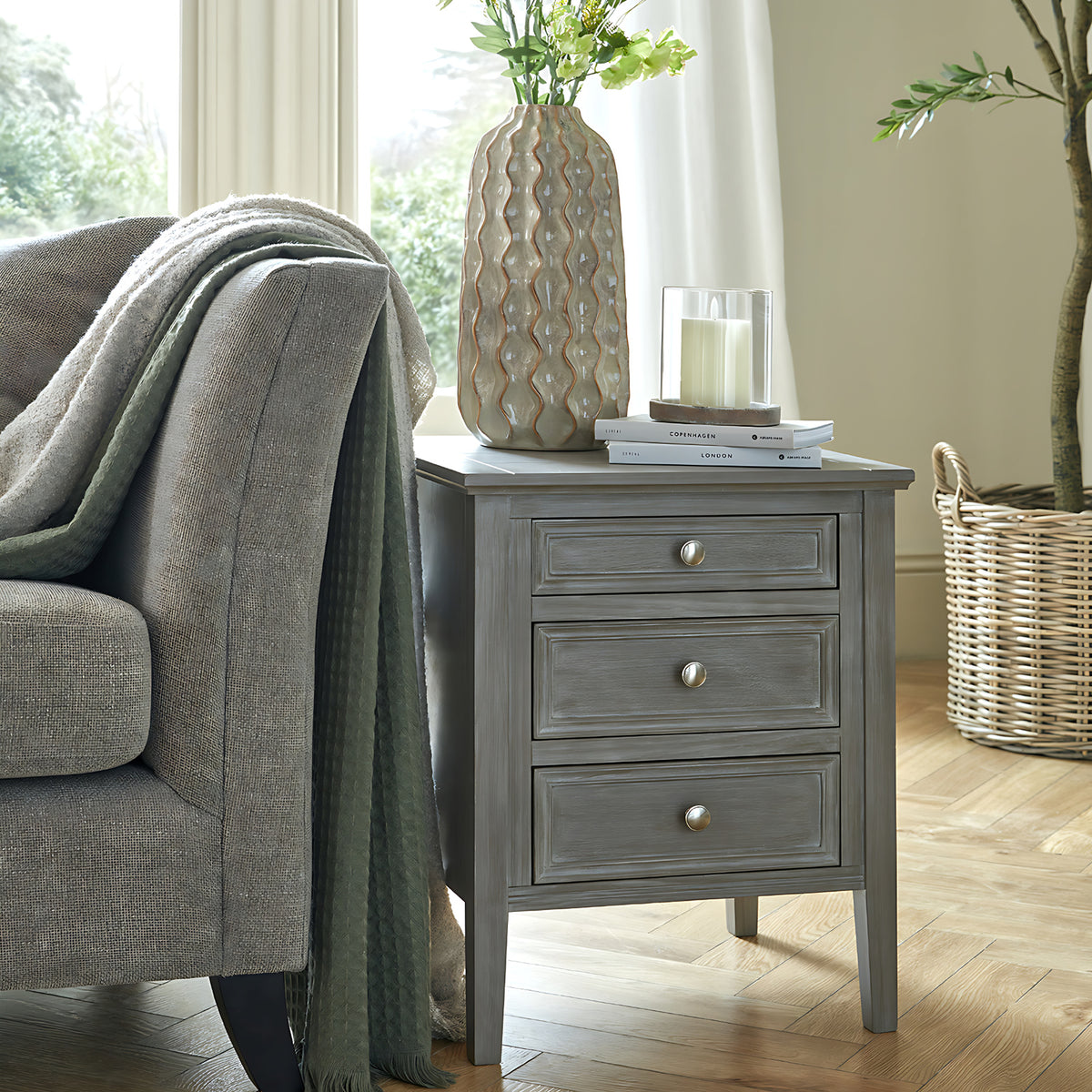 Grey-brown Drawerframe Bedside Table beside a grey armchair, with three framed drawers and a vase, books and glass candle holder on top.