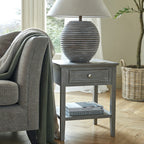 Drawerframe Side Table beside a grey armchair, styled with a large ribbed lamp on top and books on the lower shelf.