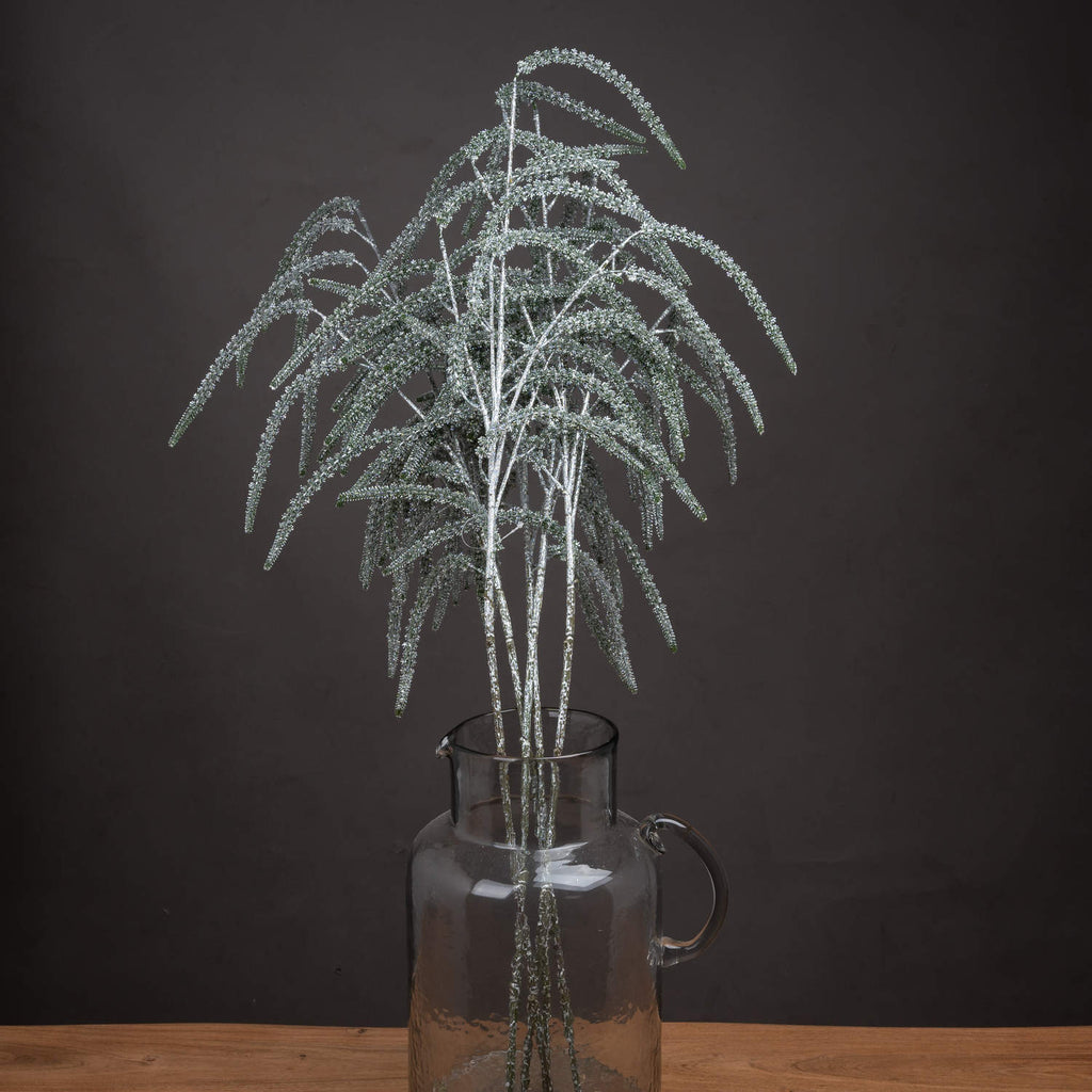 Artificial amaranthus stems in a clear handled jug on a wooden surface, their silver shimmer visible against a dark wall.