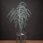 Artificial amaranthus stems in a clear handled jug on a wooden surface, their silver shimmer visible against a dark wall.