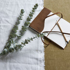 Grey-green artificial foliage branches laid diagonally across white cloth beside an open brown-covered notebook and pencil on a tan woven surface.