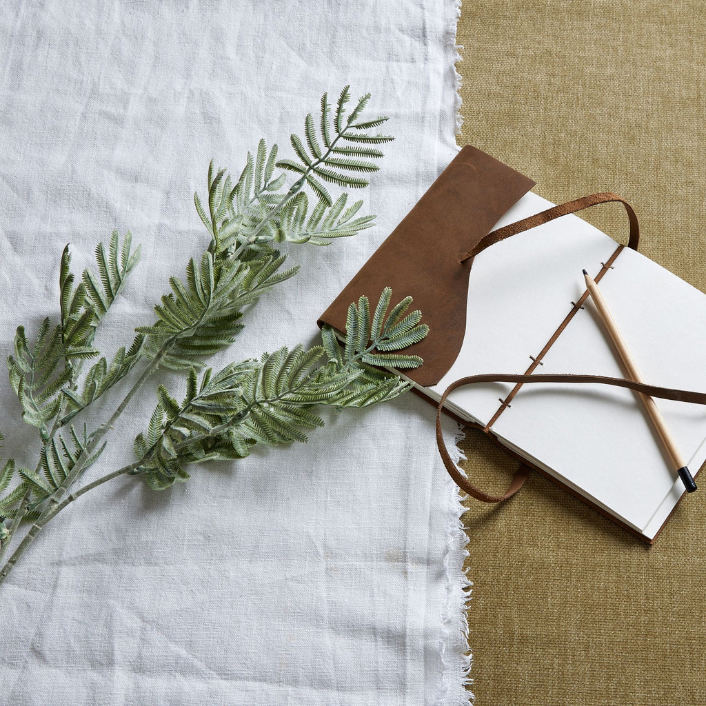 Grey-green artificial foliage laid across white fabric beside an open brown-covered notebook and pencil on a tan textured surface.