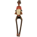 Full front view of a seated accordion musician figurine wearing a tall dark hat and red shirt, with long dangling legs and pointed shoes.