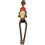 Full front view of a seated accordion musician figurine wearing a tall dark hat and red shirt, with long dangling legs and pointed shoes.