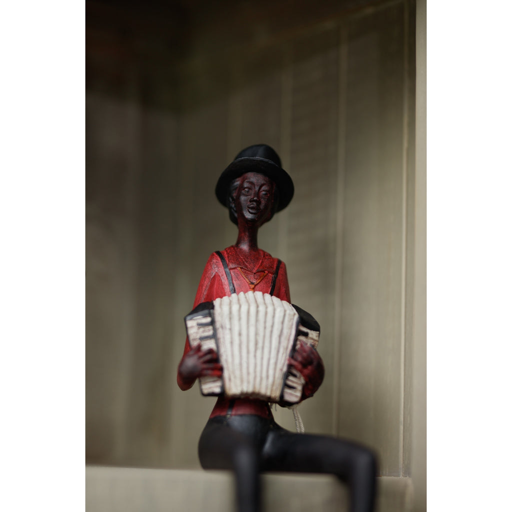 Accordion musician figurine seated on a pale shelf against panelled backing, with its legs hanging over the edge and cropped by the photograph.