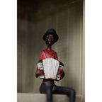 Accordion musician figurine seated on a pale shelf against panelled backing, with its legs hanging over the edge and cropped by the photograph.
