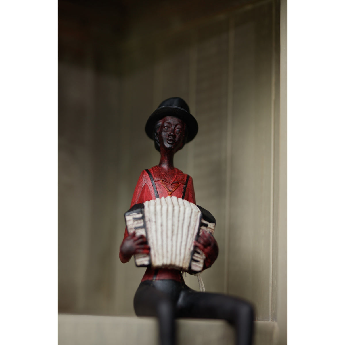 Accordion musician figurine seated on a pale shelf against panelled backing, with its legs hanging over the edge and cropped by the photograph.