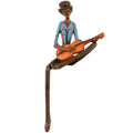 Seated guitarist figurine against a white background, wearing a tilted dark hat, blue collared shirt, reddish braces and dark tie, with an orange-brown guitar across a folded leg and the other leg hanging down.