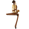 Seated saxophone player figurine on a white background, wearing a dark brimmed hat, cream shirt, orange braces and black tie, with one leg folded and the other hanging down.