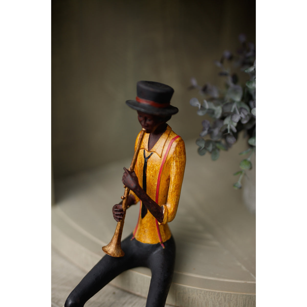 Angled close-up of the trumpeter on a pale cabinet, showing the mottled yellow shirt, reddish braces and downward-facing instrument bell.