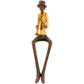 Full view of the seated trumpeter figurine against white, with a yellow shirt, reddish braces and long dark legs ending in pointed shoes.