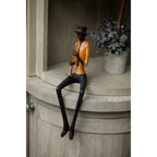 Trumpeter figurine seated on a pale wooden cabinet edge with its long legs hanging down, beside a pot of leafy greenery.