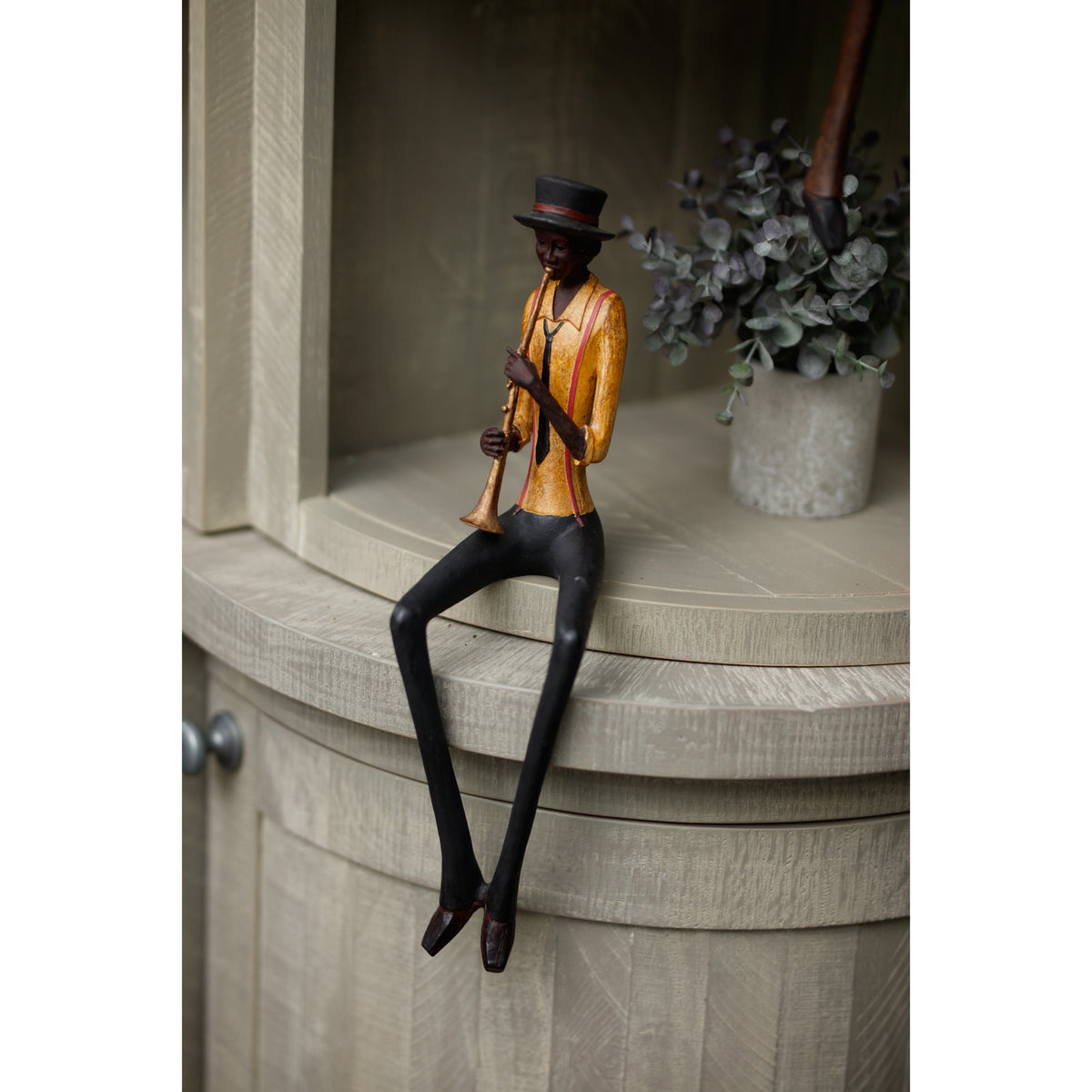 Trumpeter figurine seated on a pale wooden cabinet edge with its long legs hanging down, beside a pot of leafy greenery.