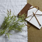 Artificial budded foliage laid diagonally across white fringed cloth beside an open brown-covered notebook and pencil on a tan woven surface.