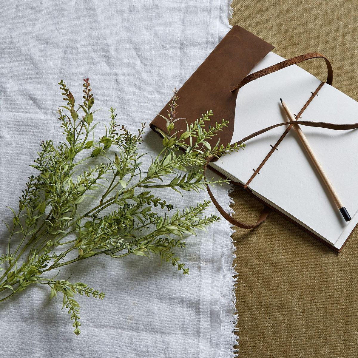 Artificial budded foliage laid diagonally across white fringed cloth beside an open brown-covered notebook and pencil on a tan woven surface.