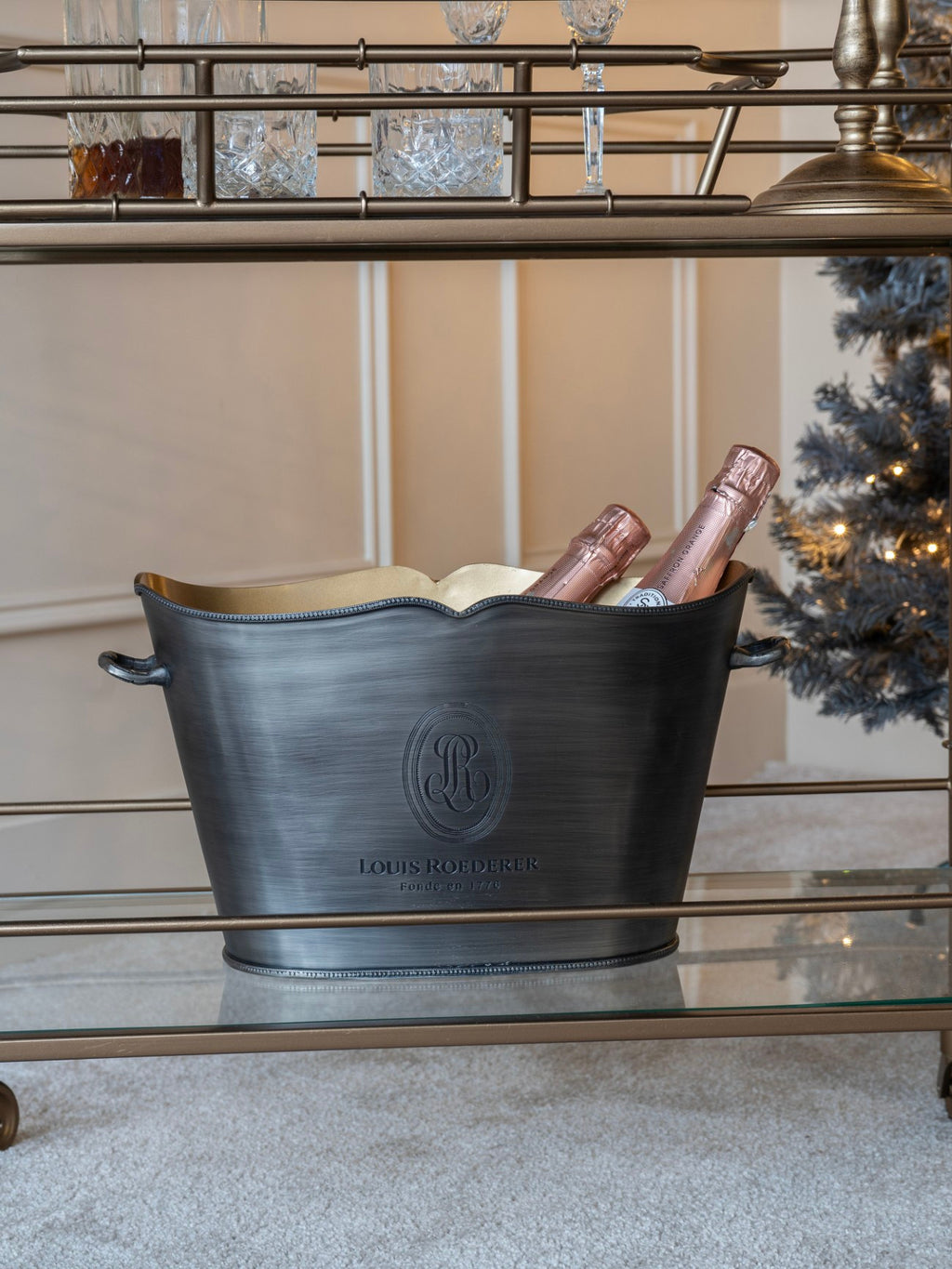 Oval champagne bucket holding two bottles on the lower glass shelf of a drinks trolley, beside a lit Christmas tree.