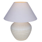 Table lamp switched on, with a warm glow visible at the top of the pale woven shade above the banded ceramic base.