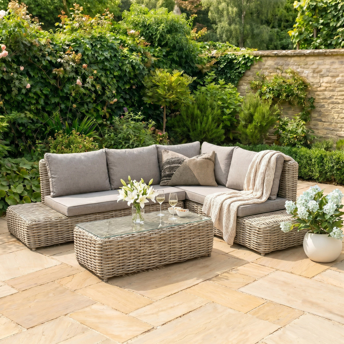 Woven L-shaped corner seating shown with pale cushions and extended end platforms on a stone patio, styled with a cream throw and a transparent-topped table amid garden greenery.