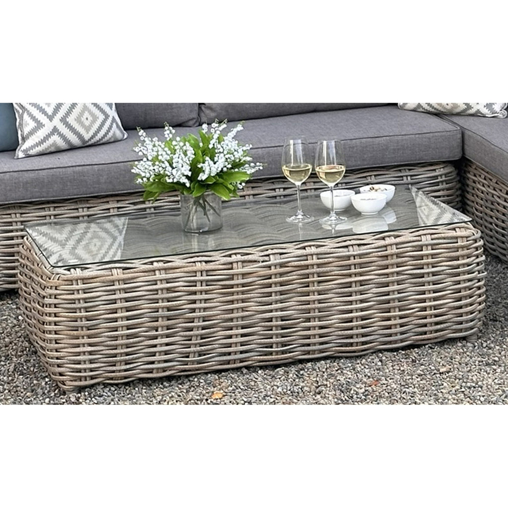 Close view of the photographed rectangular table with a transparent-looking top and rounded woven base, styled with white flowers, two stemmed glasses and small bowls in front of the seating.