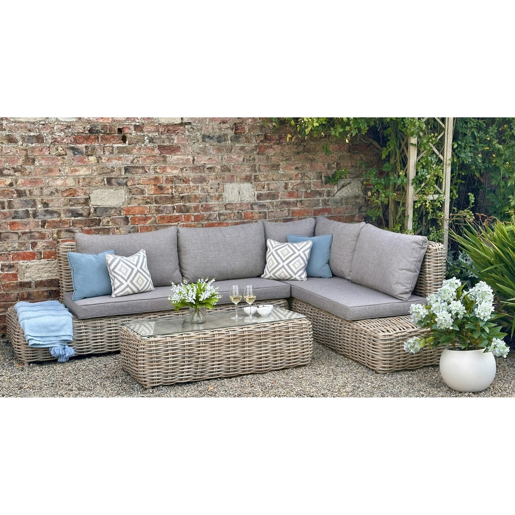 Corner seating against a brick wall, shown with grey seat and back cushions, blue and geometric scatter cushions, a blue throw on the left platform and a woven table in front.