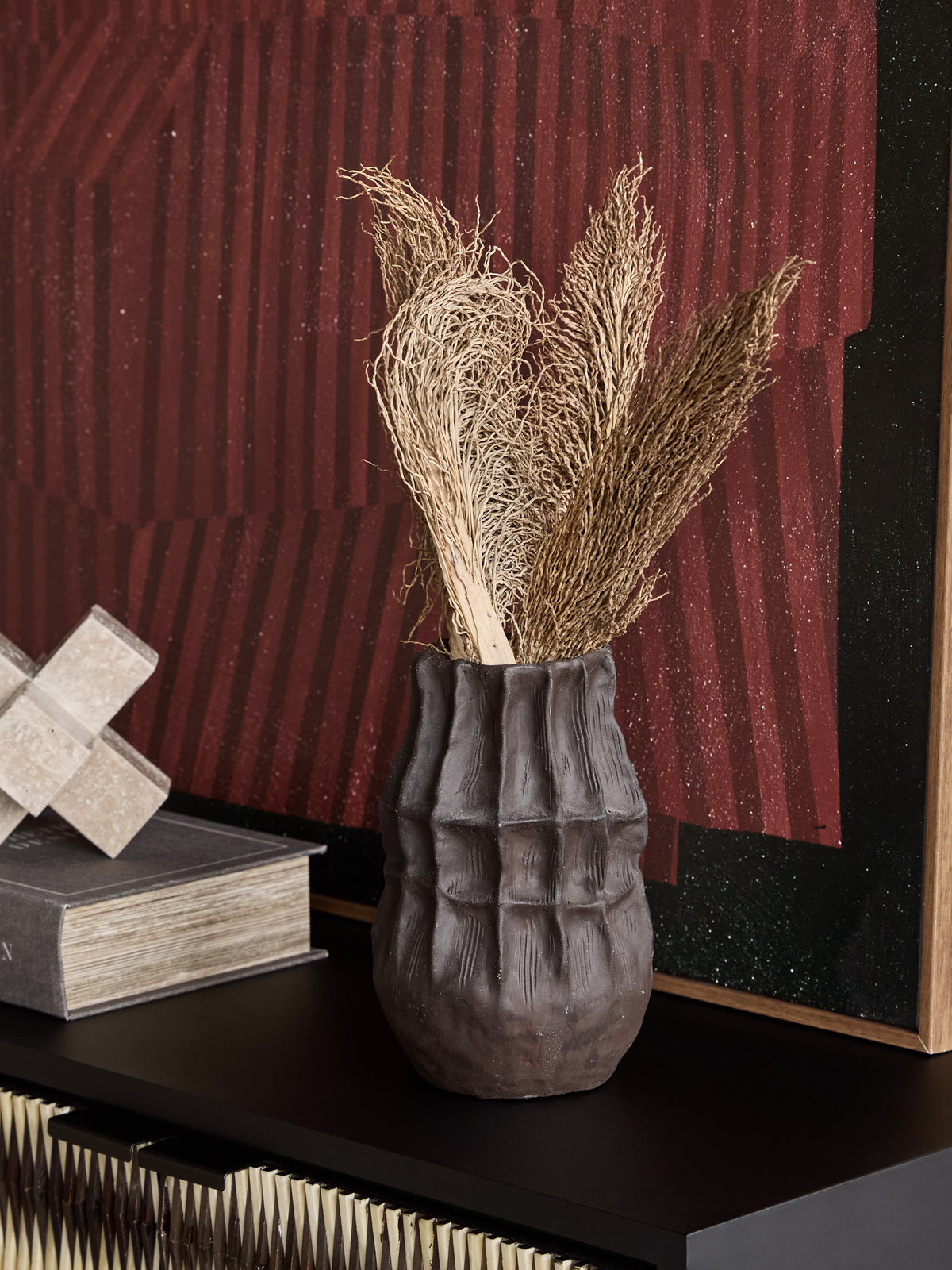 Brown faceted vase holding pale dried stems on a dark cabinet beside a book and stone ornament, with red artwork behind.