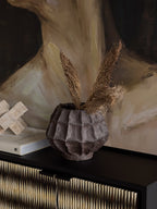Faceted brown vase filled with dried stems on a dark sideboard, beside books and a pale geometric ornament beneath abstract artwork.