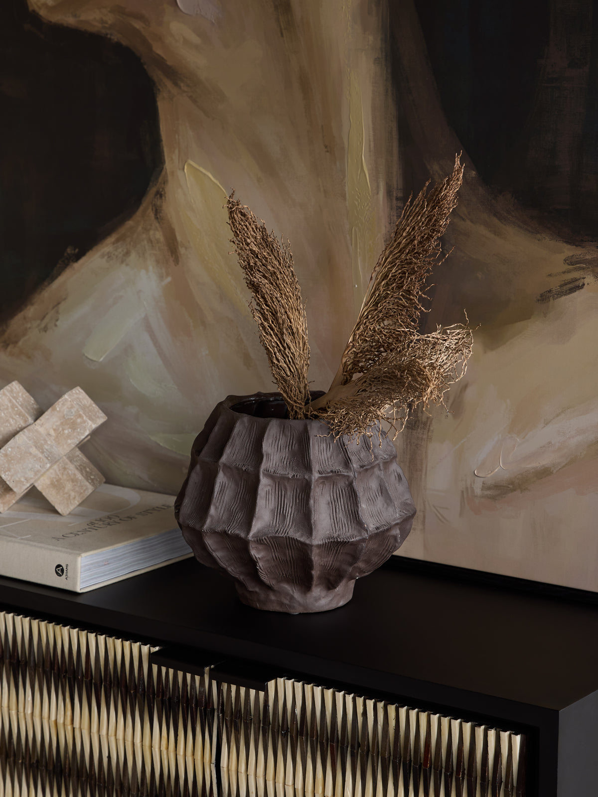 Faceted brown vase filled with dried stems on a dark sideboard, beside books and a pale geometric ornament beneath abstract artwork.