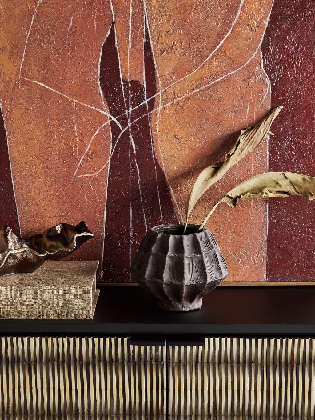 Brown faceted vase holding two dried leaves on a dark console beside stacked books, with warm abstract artwork behind.