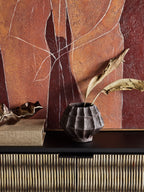 Brown faceted vase holding two dried leaves on a dark console beside stacked books, with warm abstract artwork behind.