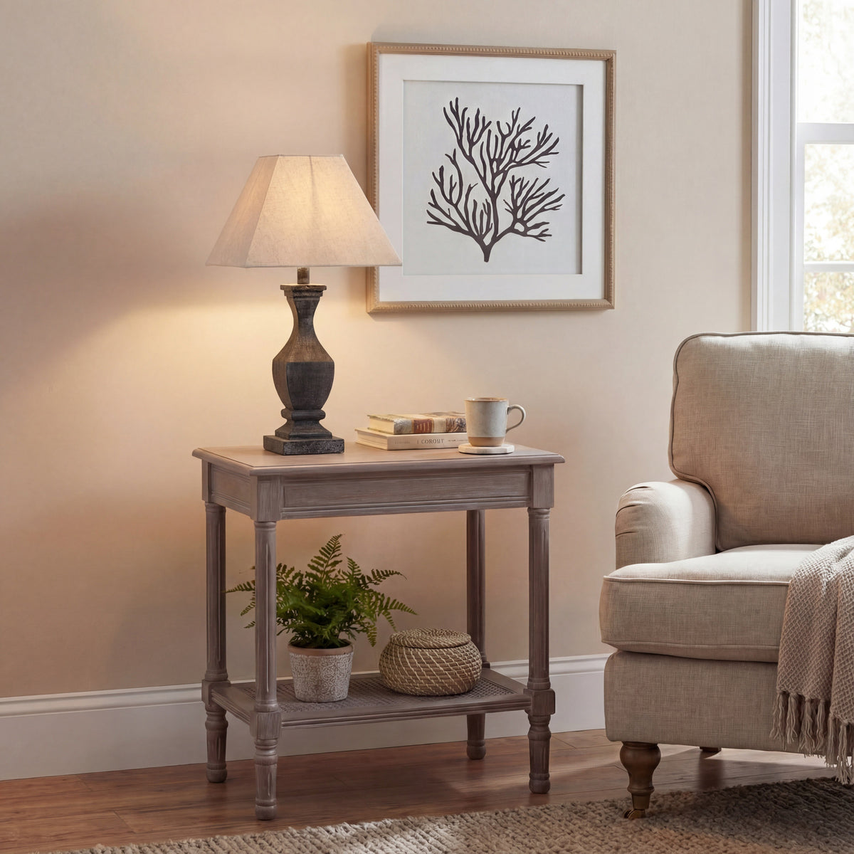 Table lamp with a glowing pale shade on a side table beside books and a mug, next to a cream armchair beneath a framed branching print.