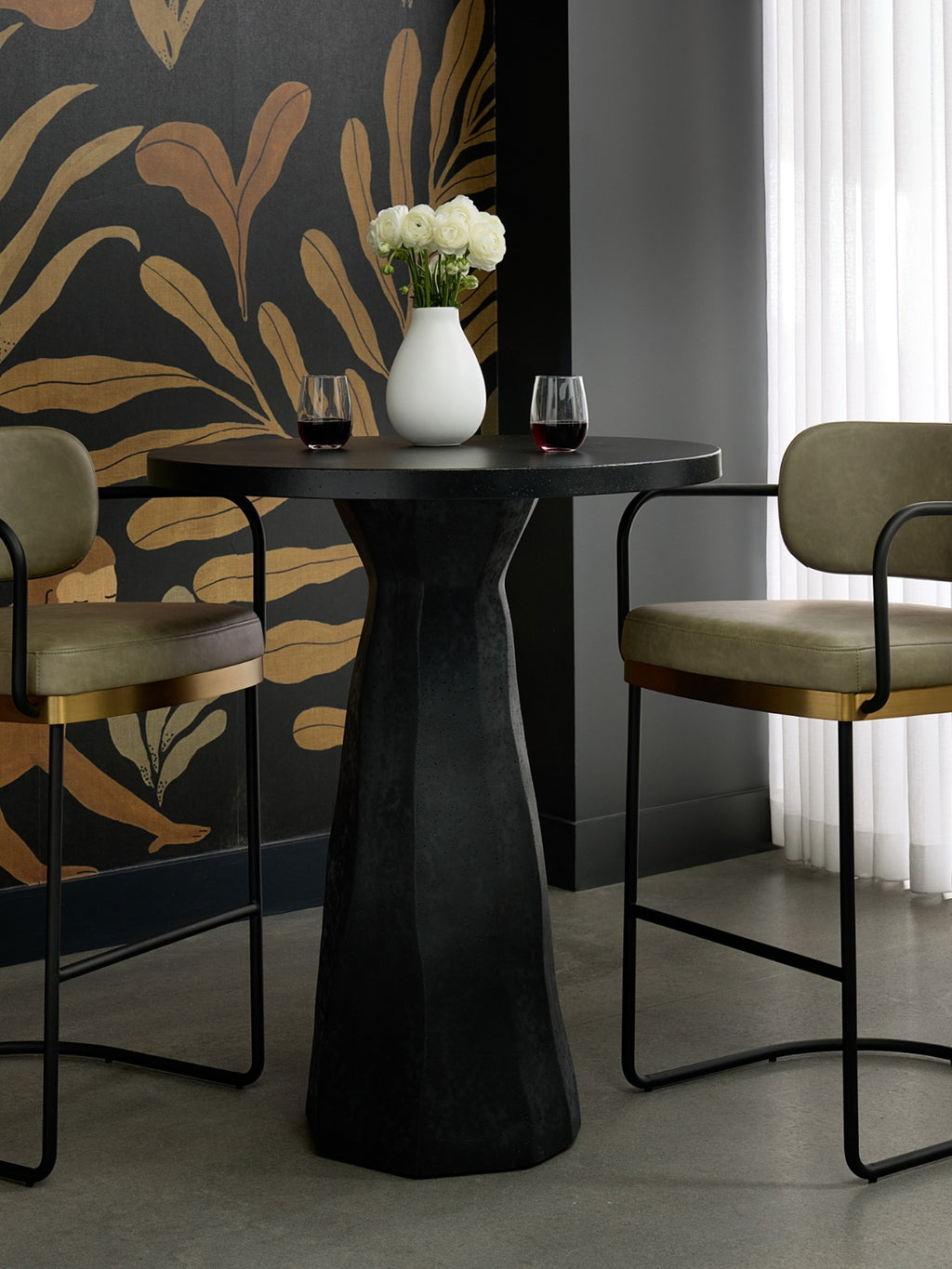 Black pedestal bar table between two olive upholstered stools, with wine glasses and white flowers against gold leaf-patterned wallpaper.