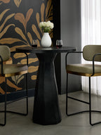 Black pedestal bar table between two olive upholstered stools, with wine glasses and white flowers against gold leaf-patterned wallpaper.