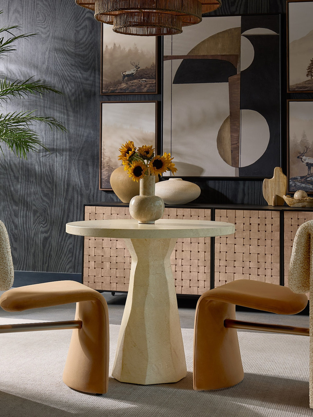 Round taupe bistro table between two tan chairs, topped with a vase of sunflowers beneath a woven pendant.