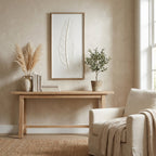 Framed feather artwork hanging above a pale console with books, a vase of dried grasses and a potted tree beside a cream armchair.
