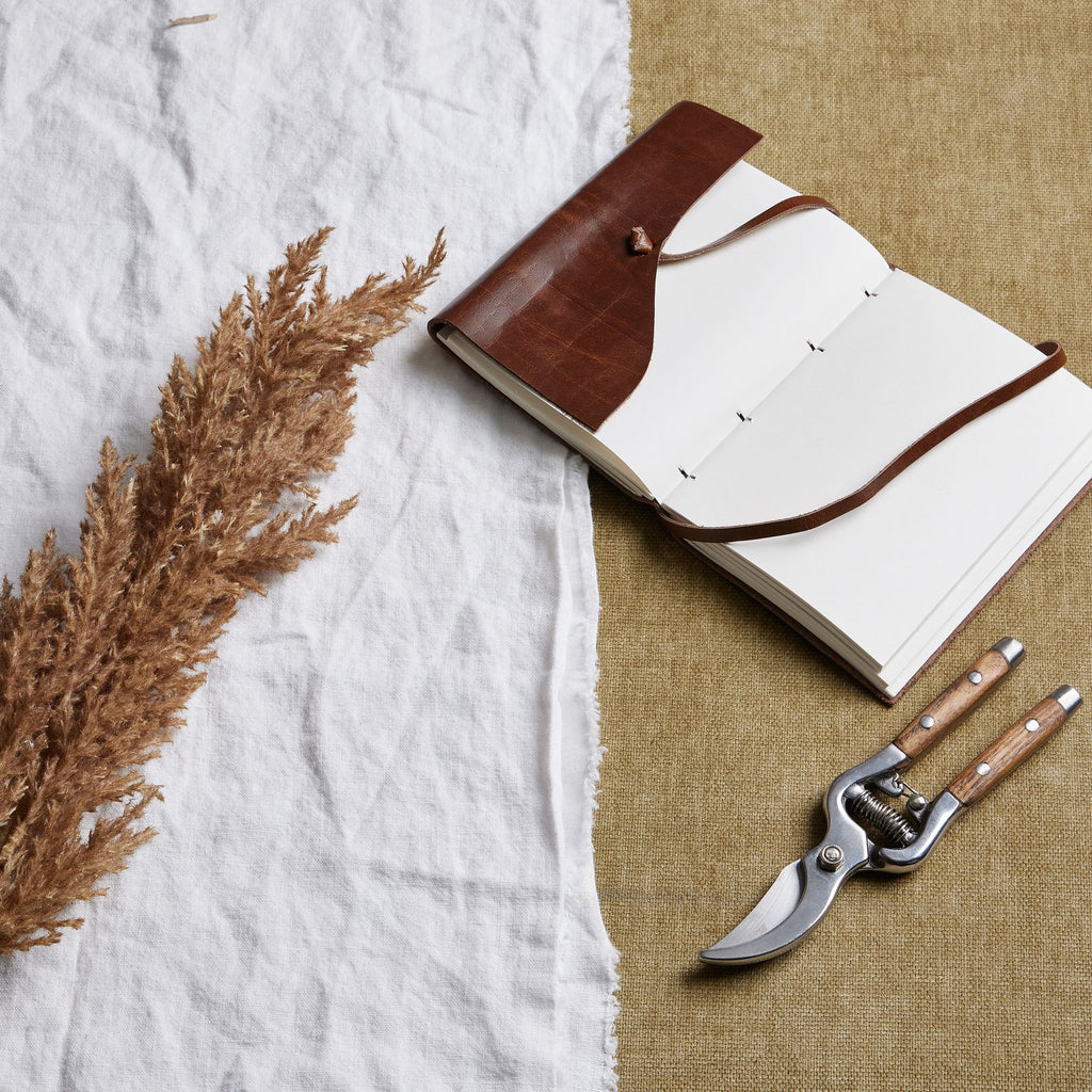 Brown feathery plume laid diagonally on white fabric beside an open brown-covered notebook and pruning shears on a tan woven surface.