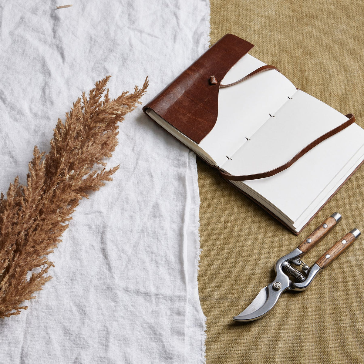 Brown feathery plume laid diagonally on white fabric beside an open brown-covered notebook and pruning shears on a tan woven surface.
