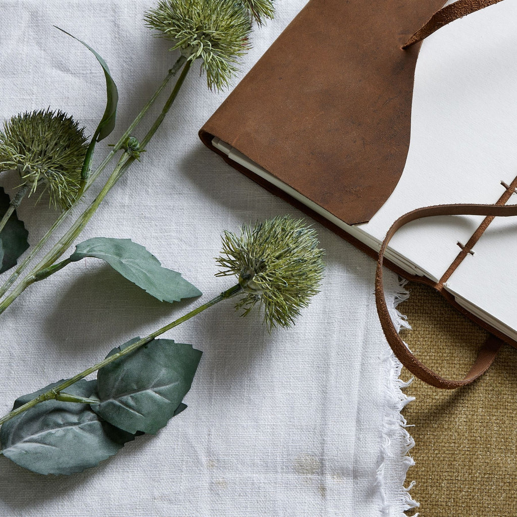 Artificial globe flower heads and green leaves laid across pale woven fabric beside an open notebook with a brown cover and wraparound tie.