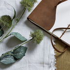 Artificial globe flower heads and green leaves laid across pale woven fabric beside an open notebook with a brown cover and wraparound tie.
