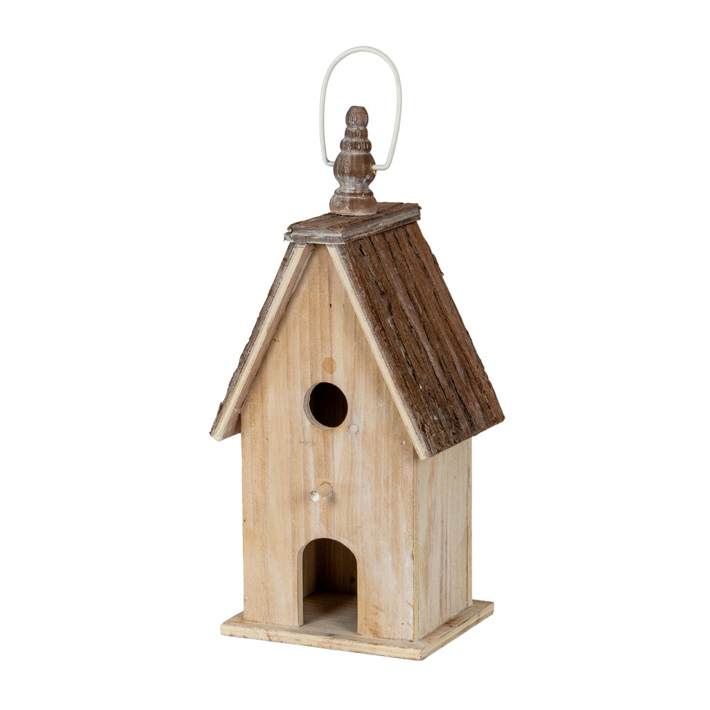 Three-quarter front view of the birdhouse, showing its pale washed body, dark pitched roof, square plinth and finial beneath a pale loop.