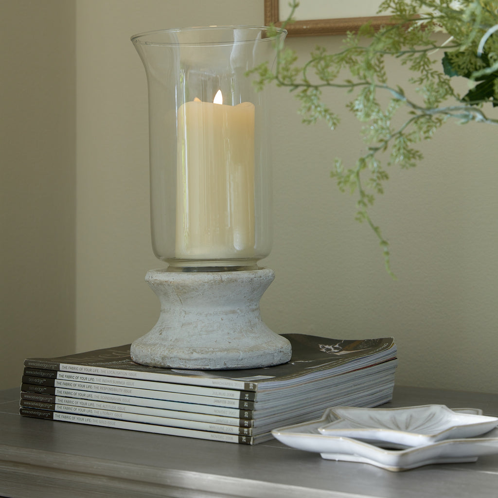 Lantern shown with a cream candle inside the clear sleeve, standing on stacked magazines beside white leaf-shaped dishes, with greenery and a framed mirror behind.
