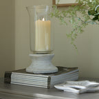 Lantern shown with a cream candle inside the clear sleeve, standing on stacked magazines beside white leaf-shaped dishes, with greenery and a framed mirror behind.