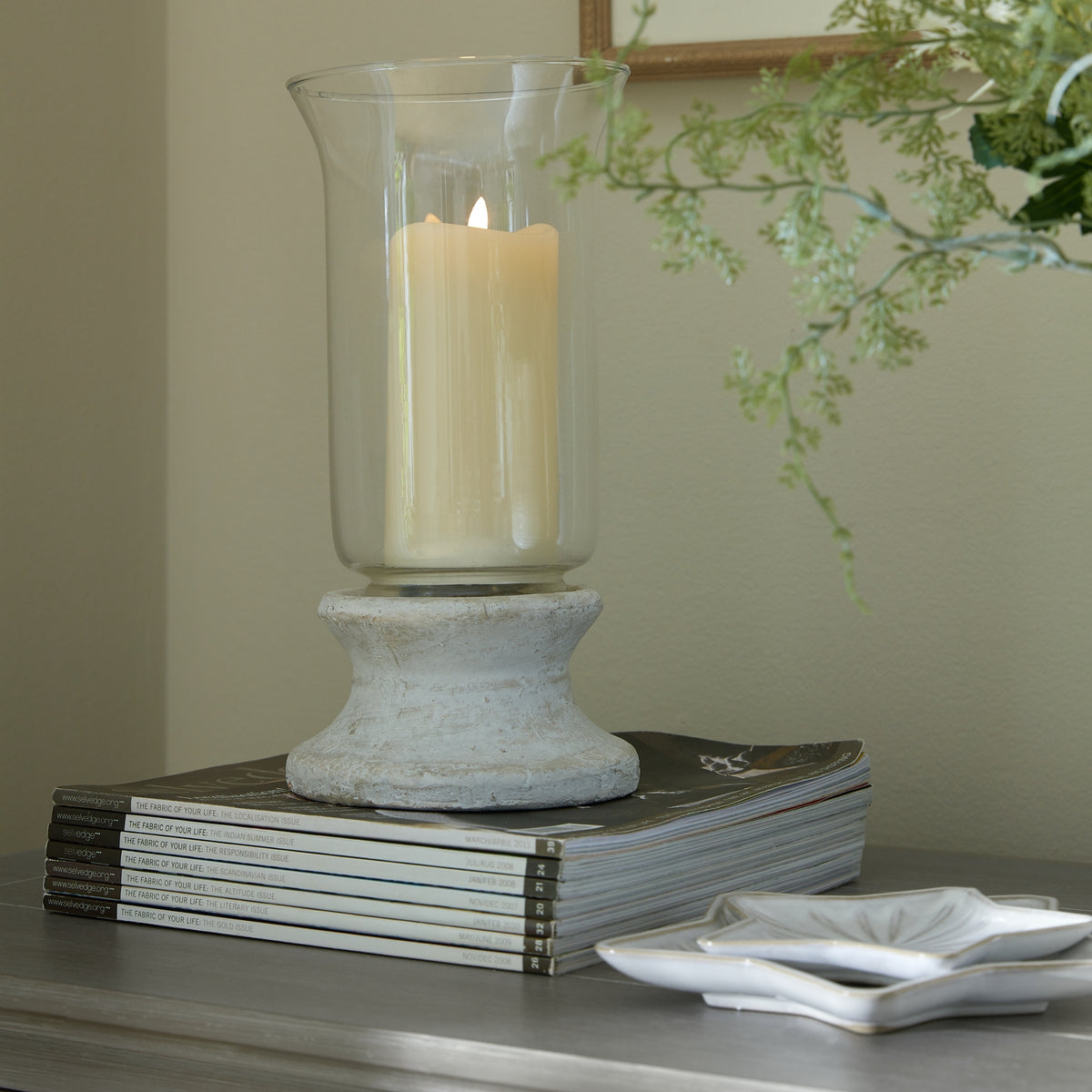 Lantern shown with a cream candle inside the clear sleeve, standing on stacked magazines beside white leaf-shaped dishes, with greenery and a framed mirror behind.
