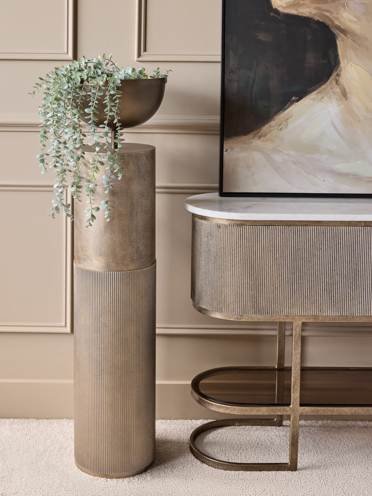 Gold pedestal planter filled with trailing green foliage beside a fluted console and abstract artwork against a cream panelled wall.
